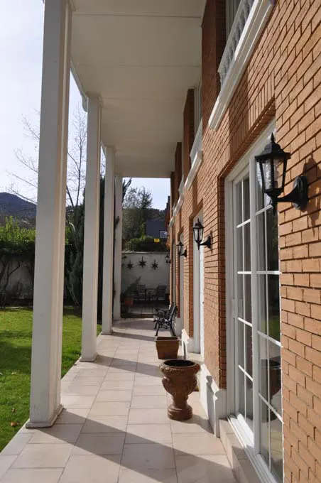 Covered patio with white columns, brick wall, and tiled floor. Black sconces and white-framed windows line the wall. Green lawn visible.