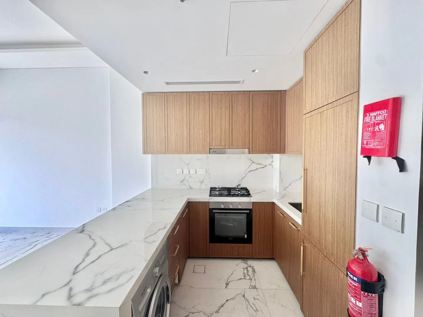 A modern kitchen with light wood cabinets, white marble countertops and backsplash, and stainless steel appliances. A red fire blanket and extinguisher are mounted on the wall.
