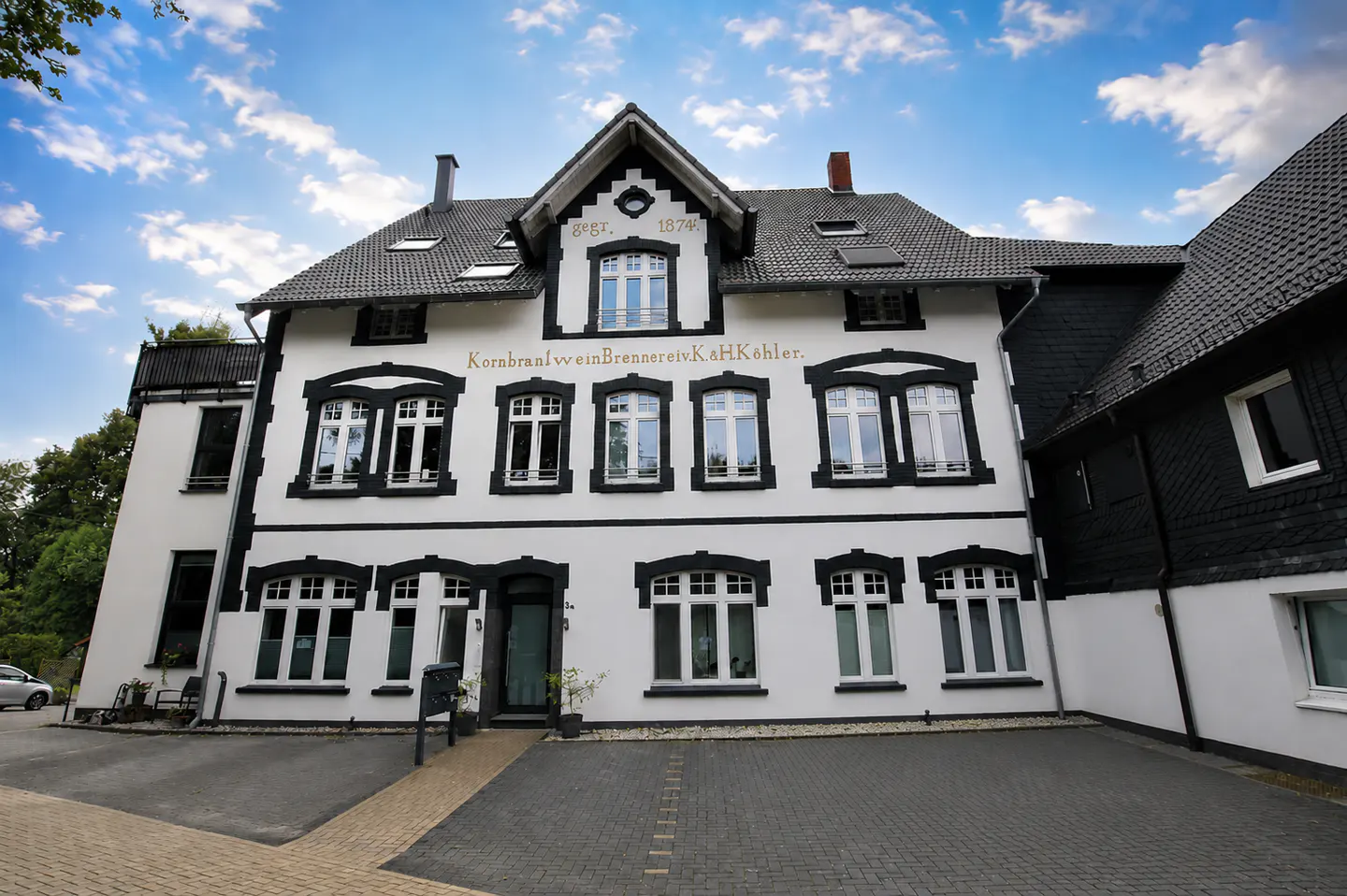 Two-story white building with black trim and a gray roof under a blue sky. "KornbraalweinBrennereiK.a.H.Köhler" is written on the building.
