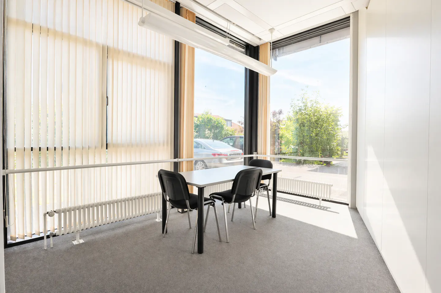 Bright office space with a white table and four black chairs. Large windows with vertical blinds offer a view of trees and a car.
