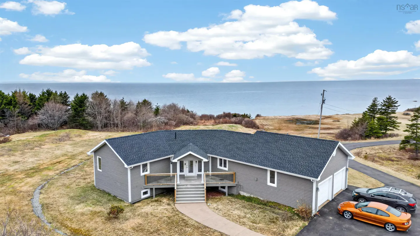 Gray house with a dark roof, front deck, and two-car garage. Two cars are parked in the driveway. Ocean and trees in the background.