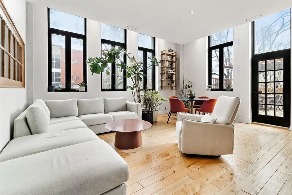 Bright living room with a white sectional sofa, a round coffee table, and a white armchair on a wooden floor. Tall windows with black frames.