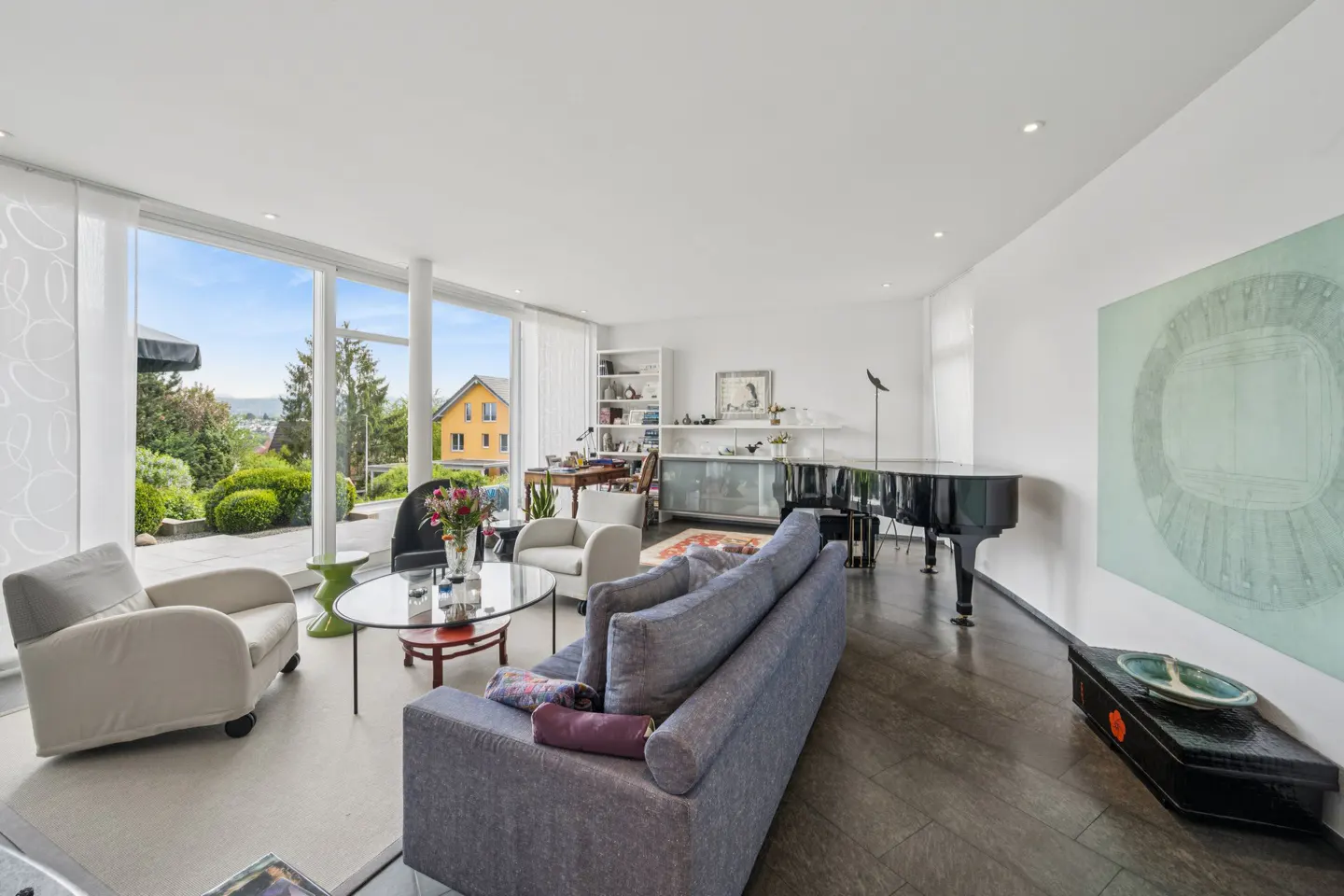 Bright living room with a gray sofa, armchairs, a black piano, and a large window overlooking a garden.