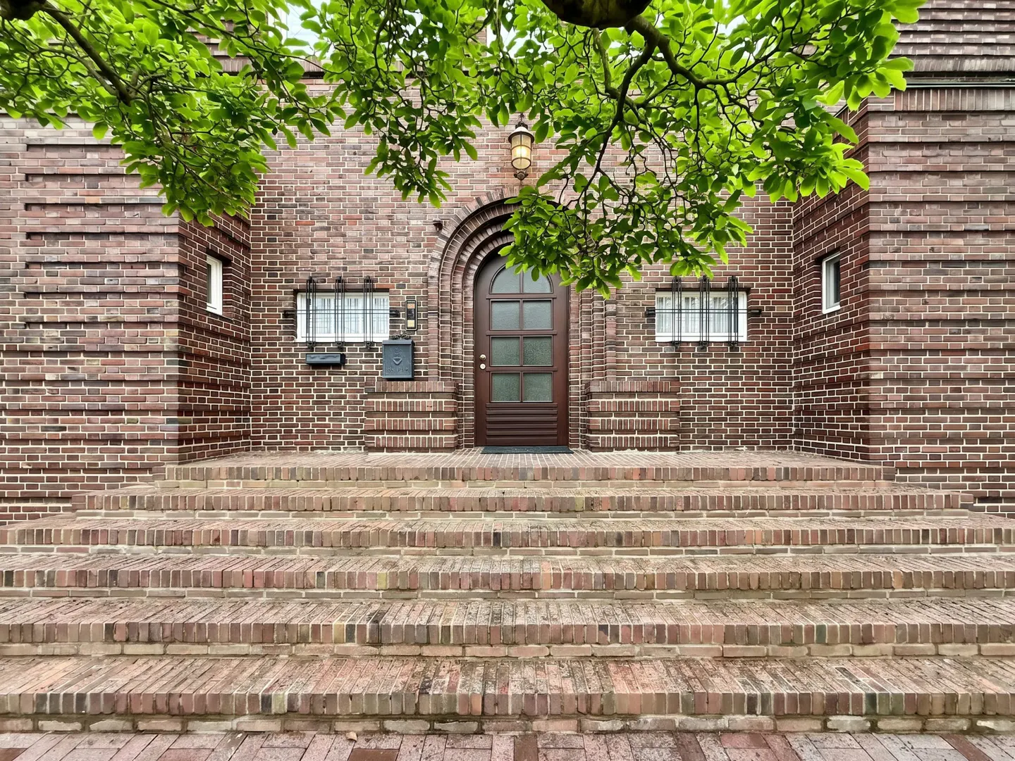Brick house with steps leading to a brown door under an arched entryway. Green leaves frame the top of the image.