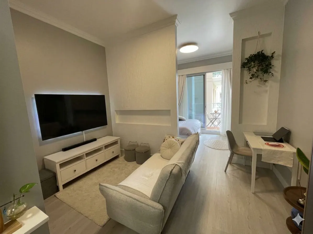 A bright, modern apartment interior with a TV, sofa, desk, and a glimpse of a bedroom and balcony.