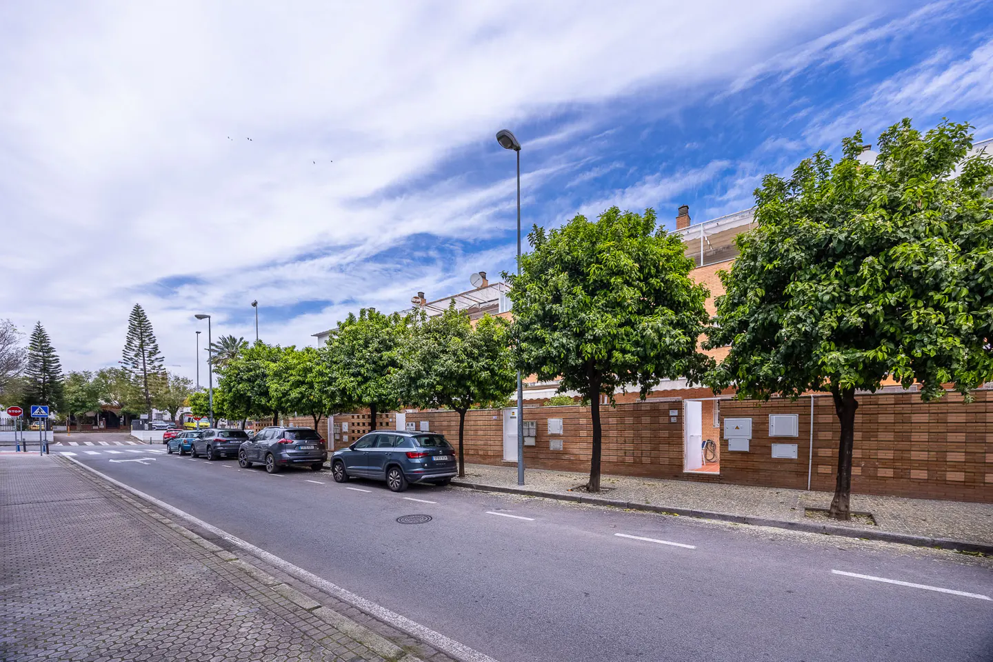 Street view of parked cars along a brick building with green trees under a blue, cloudy sky.