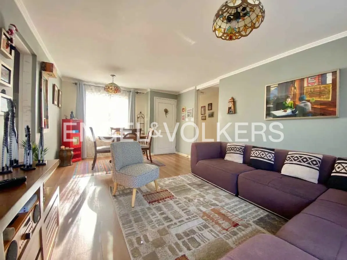 A living room with a purple sofa, patterned rug, and dining table. A gray chair sits near the rug. A painting hangs on the wall.