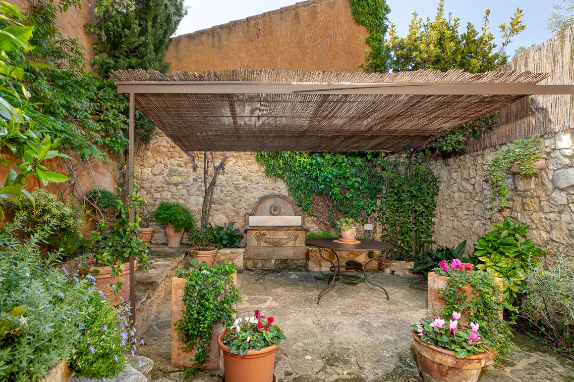 Outdoor patio with stone walls, a fountain, and a pergola covered with reeds. Potted plants add greenery and color.