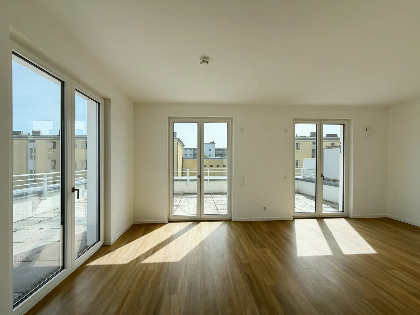 Bright, empty room with light wood floors and white walls. Glass doors lead to balconies with city views. Sunlight streams in.