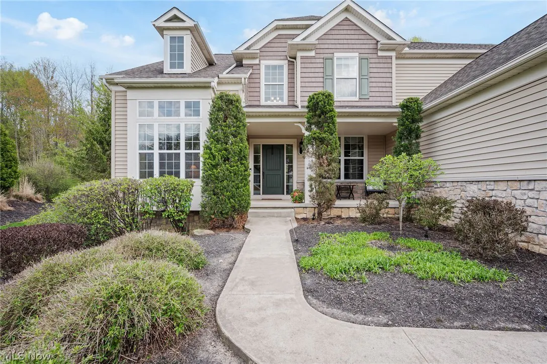 Two-story house with beige and brown siding, green door, and a concrete walkway leading to the entrance. Landscaped front yard with bushes and trees.