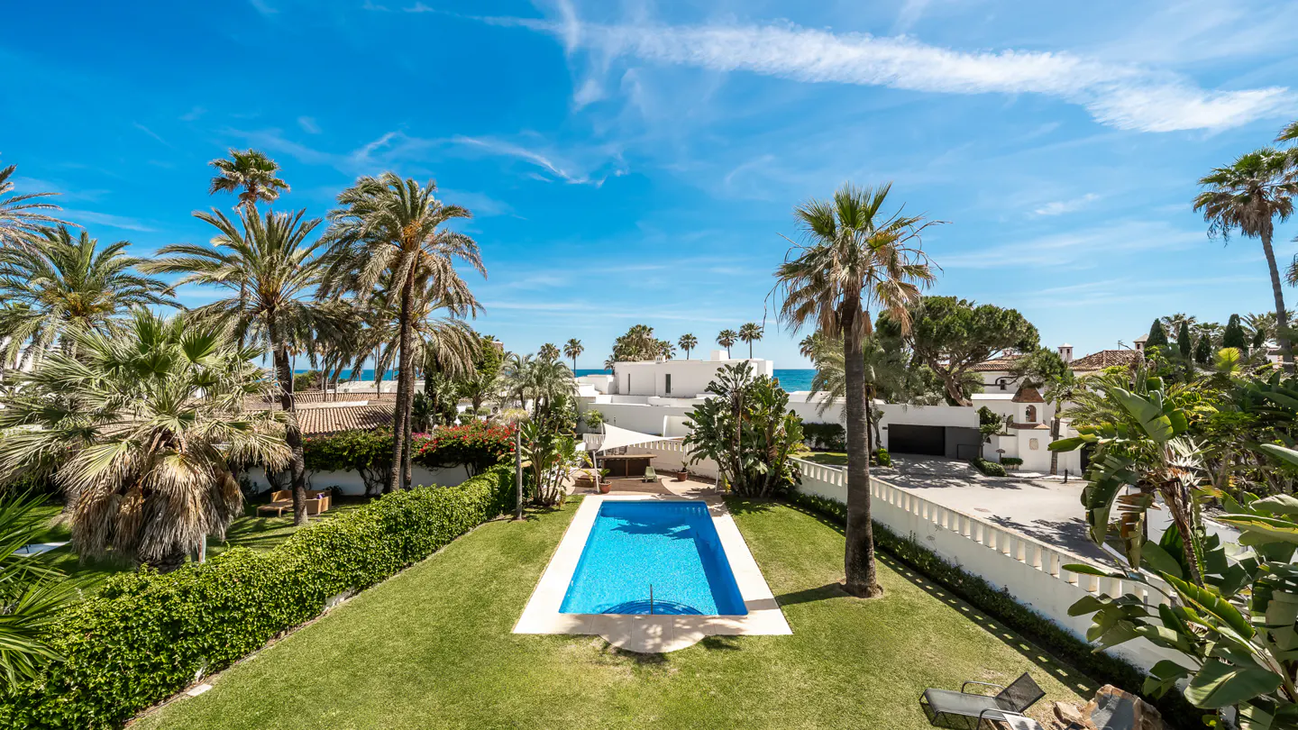 Aerial view of a rectangular blue pool surrounded by green lawn, palm trees, and a white villa under a bright blue sky.