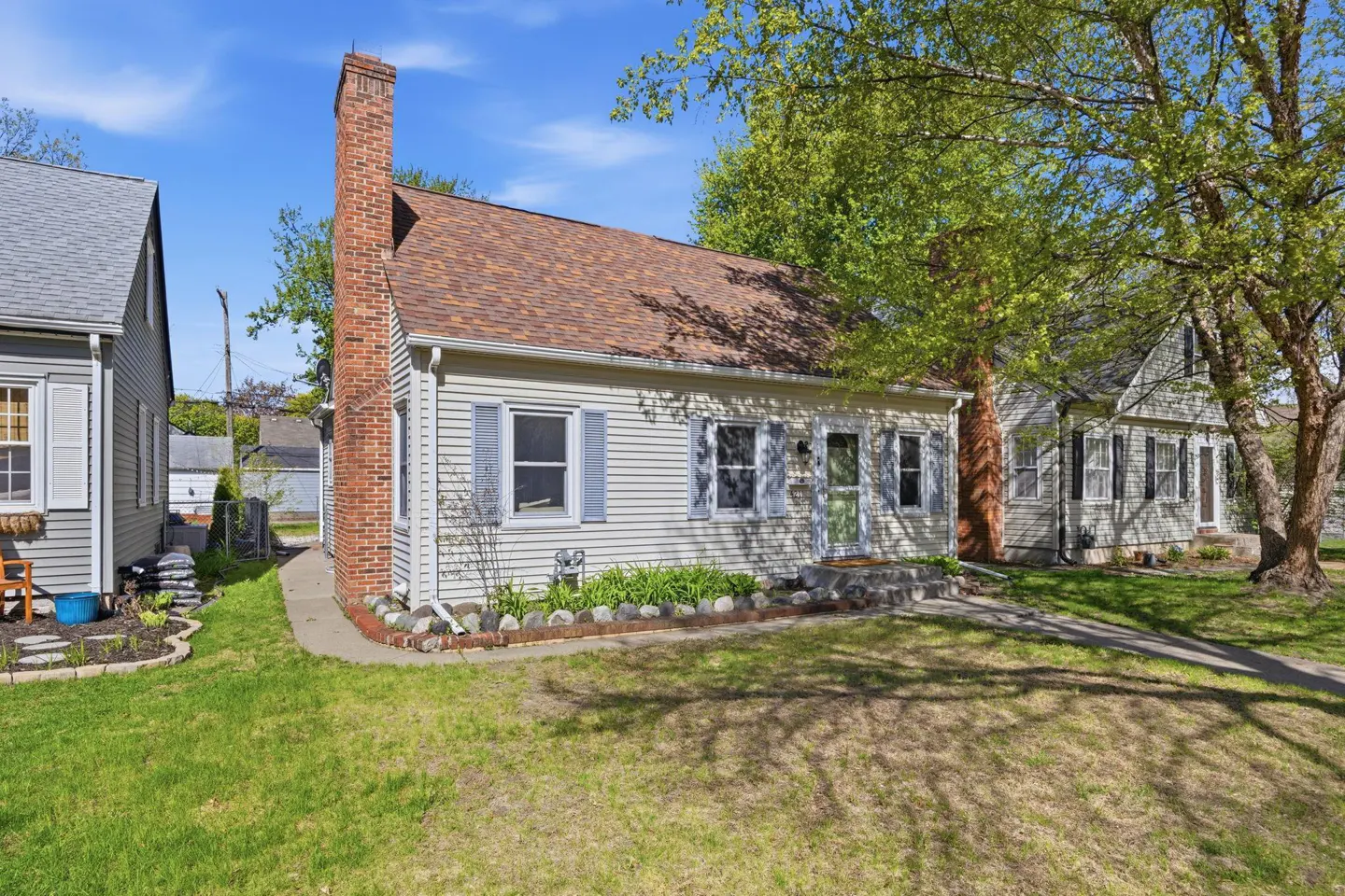 A light yellow house with a brown roof and brick chimney, surrounded by green grass and trees.