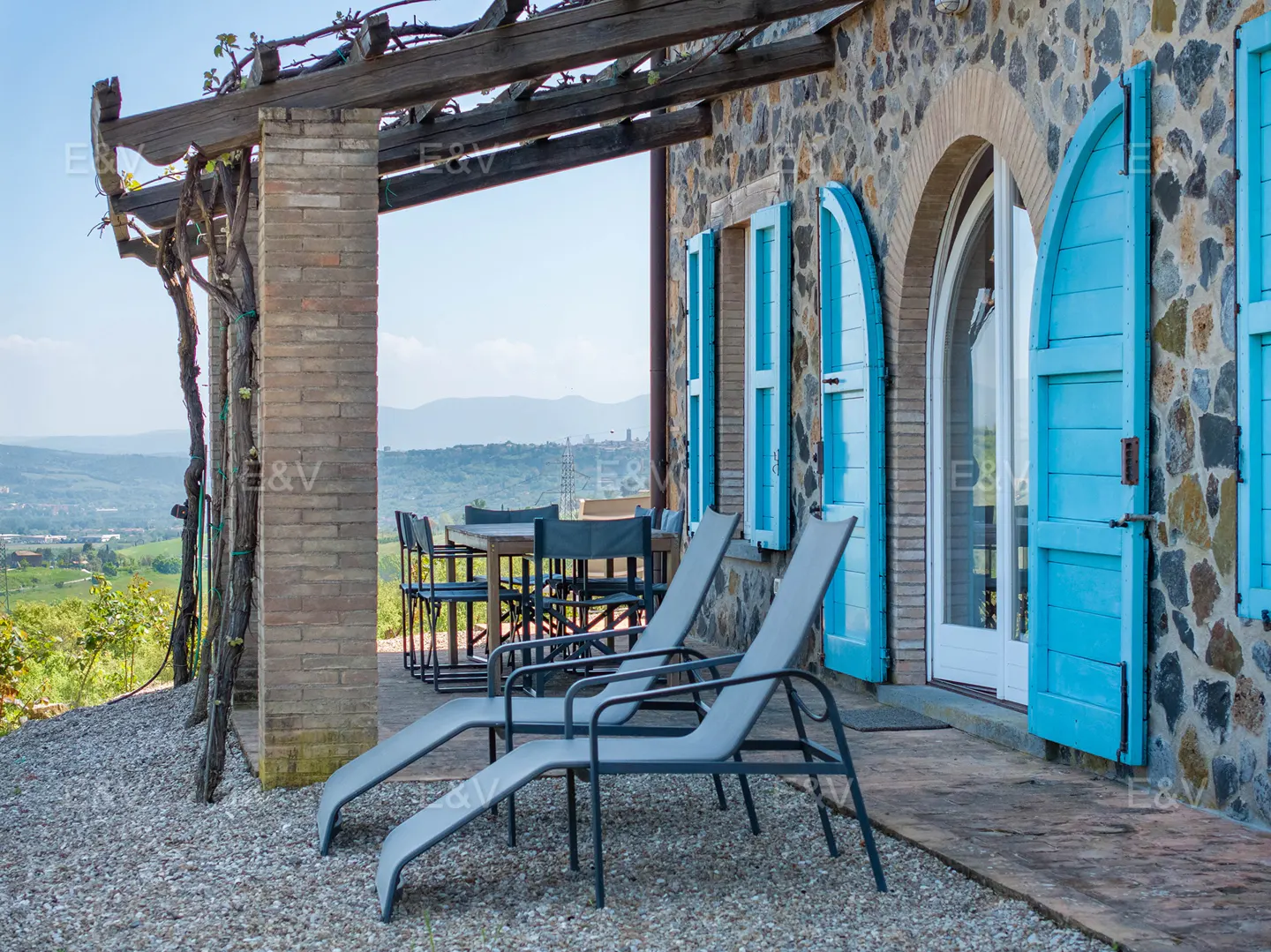 Stone house with blue shutters, two lounge chairs, and a table with chairs on a gravel patio overlooking a green landscape.