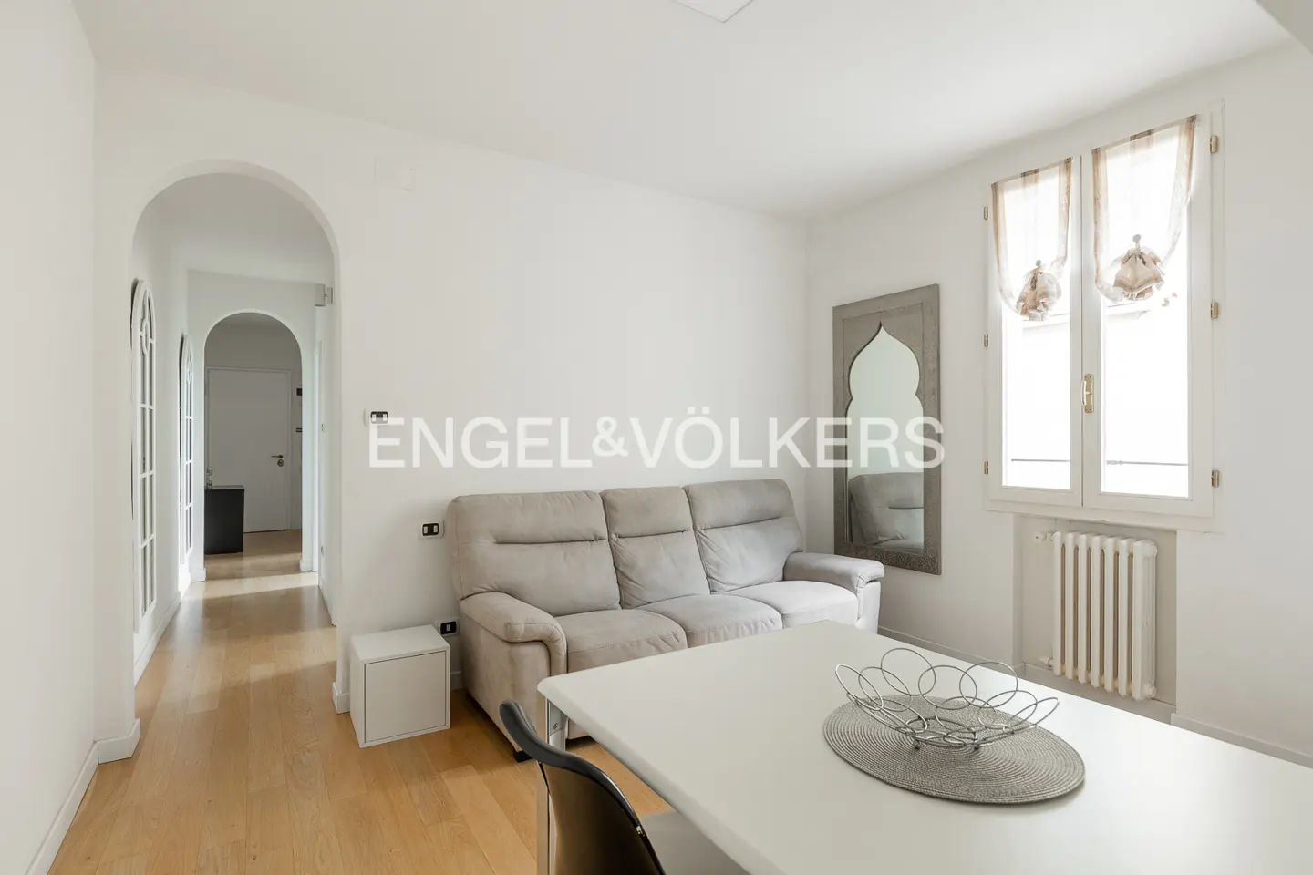 Bright living room with white walls, wood floors, and a gray sofa. A white table sits in front of the sofa, and a mirror hangs on the wall.