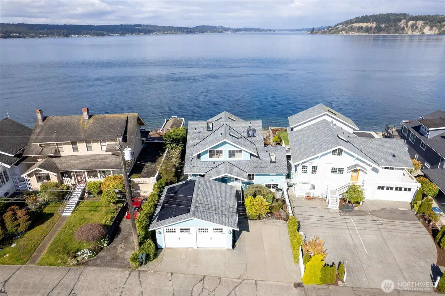 Aerial view of waterfront homes with blue water and distant land in the background. The houses are closely spaced with varying architectural styles.