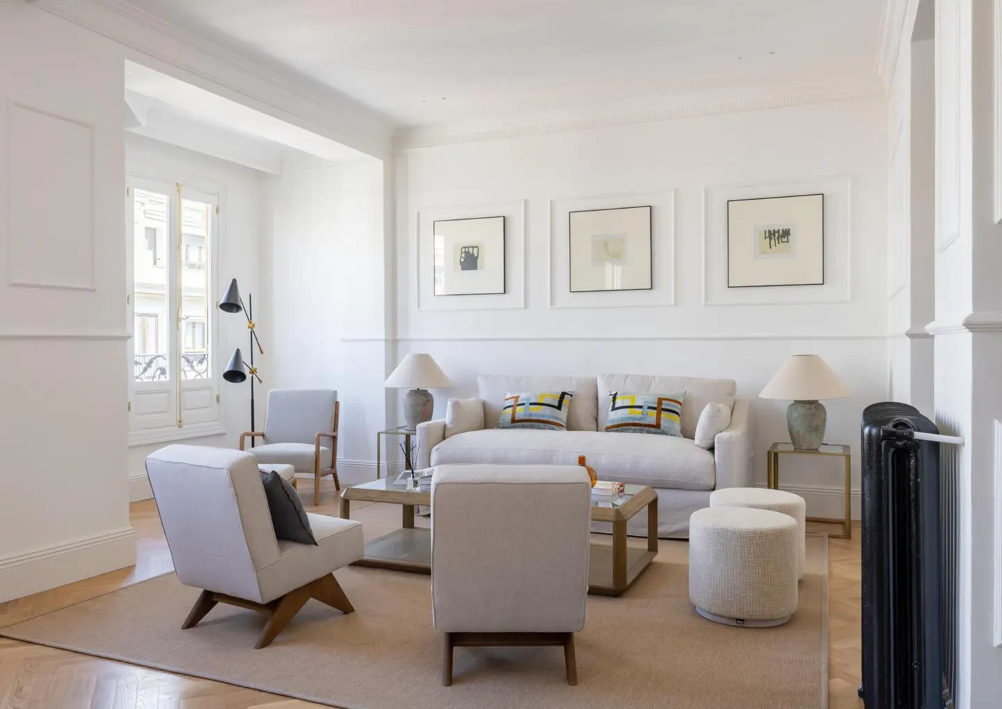 Bright living room with white walls, beige sofa and chairs, and three framed pictures above the sofa. A black floor lamp stands near a window.