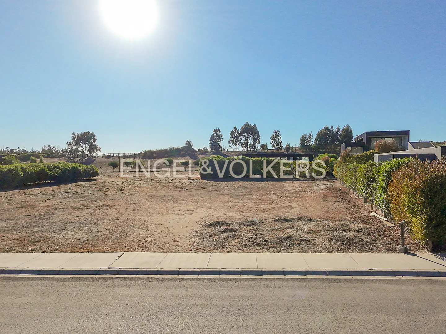Vacant lot with Engel & Völkers sign on a sunny day. Blue sky, brown dirt, and green bushes. Modern house in the background.