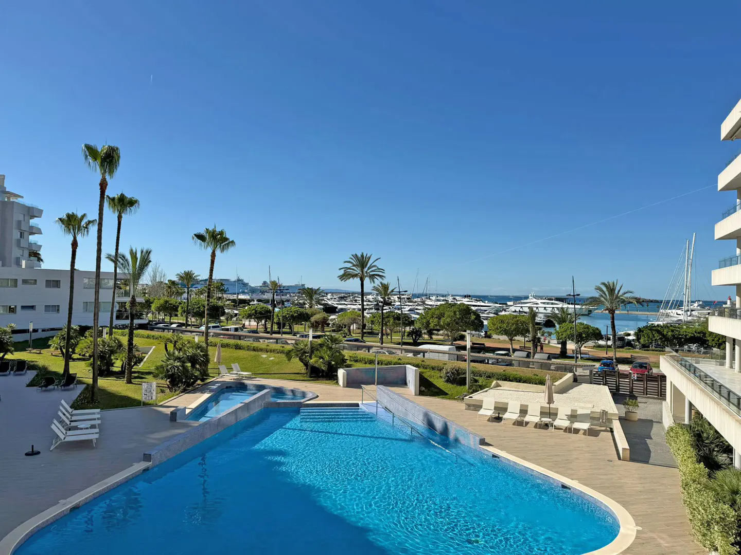 View of a blue swimming pool with palm trees, a marina with yachts, and white buildings under a clear blue sky.