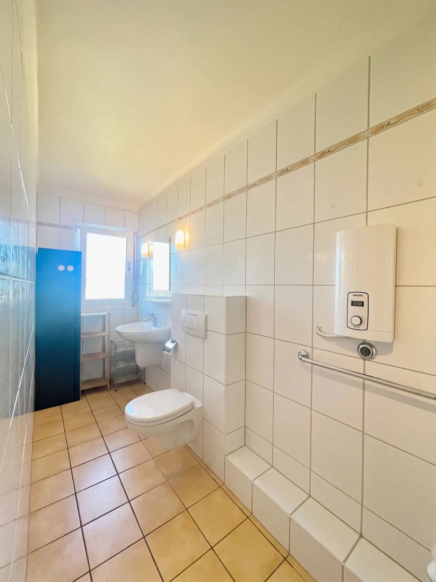 Bright bathroom with beige tile floor, white tiled walls, toilet, sink, blue cabinet, and water heater.