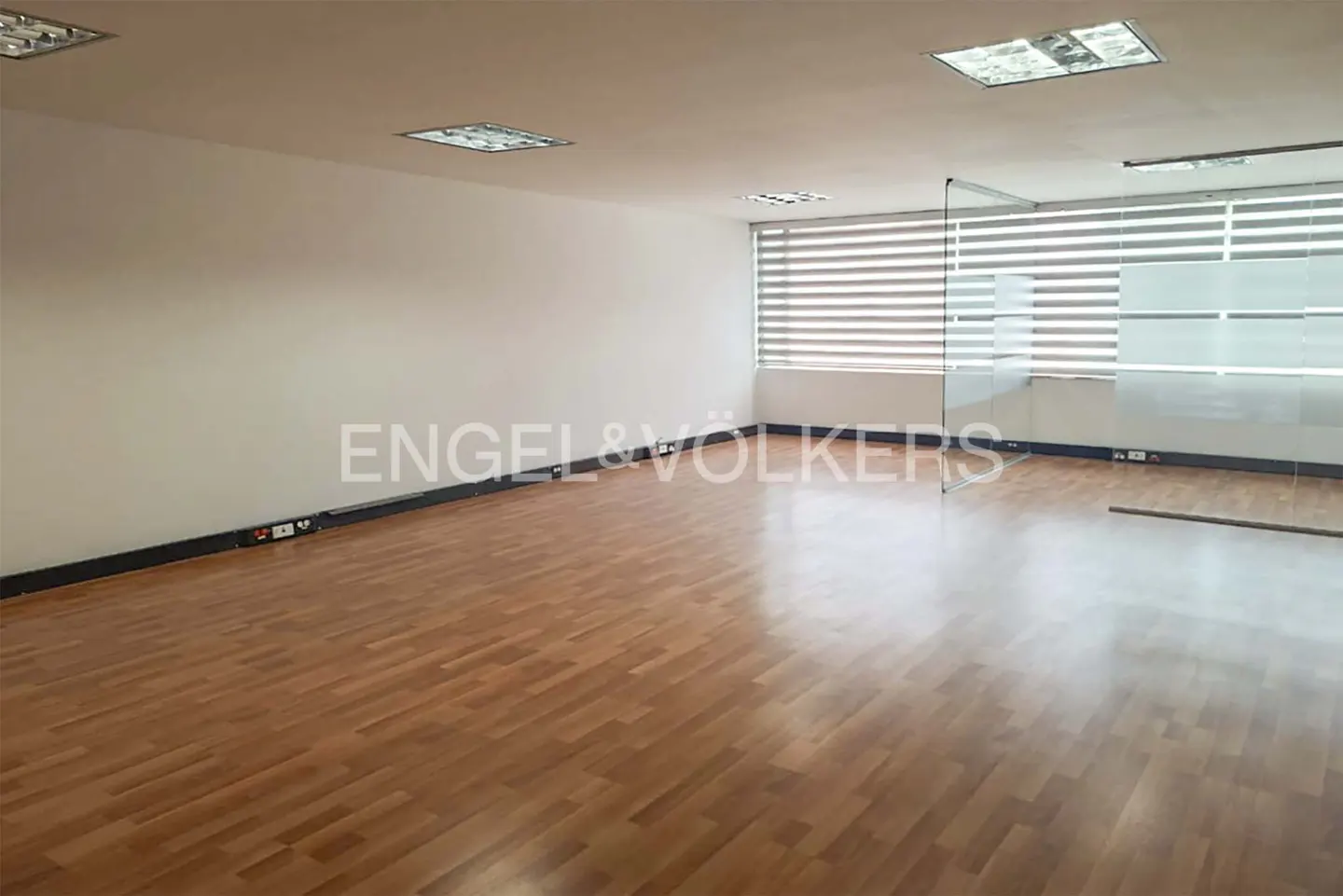 Empty office space with wood floors, white walls, and glass partitions. Blinds cover the window. Engel & Volkers logo on the wall.