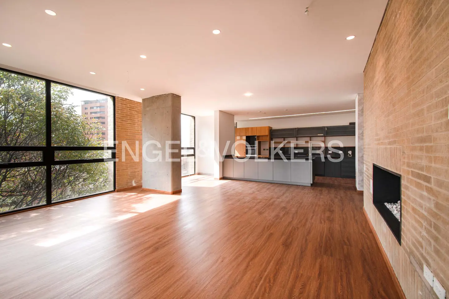 Bright, open-concept living space with wood floors, brick accent wall, and modern kitchen. Large windows offer a view of trees.