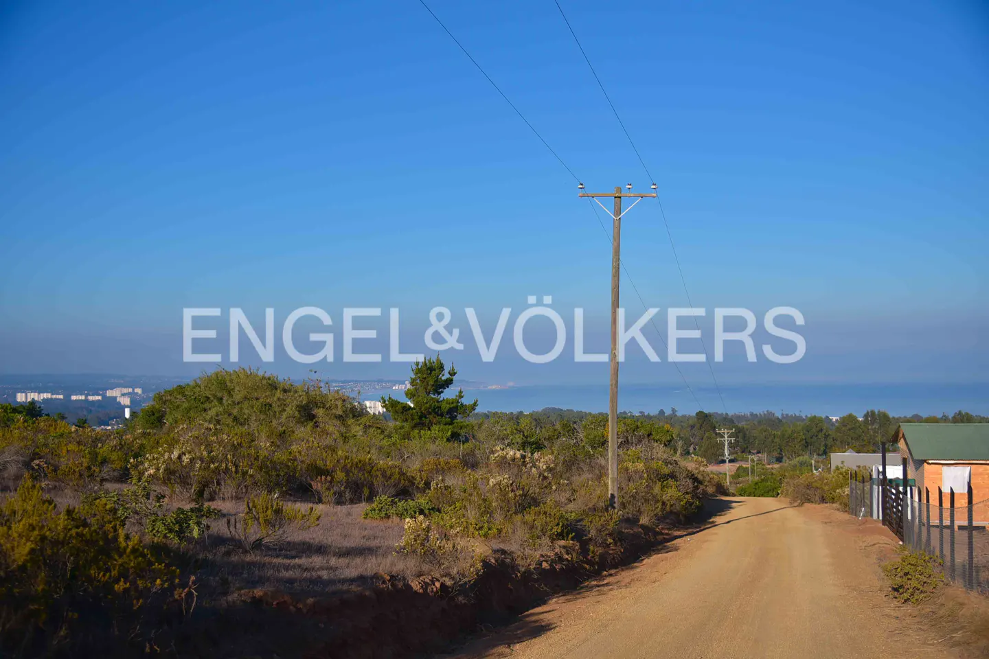 Dirt road with a utility pole on the right, leading to a house with a green roof under a clear blue sky.