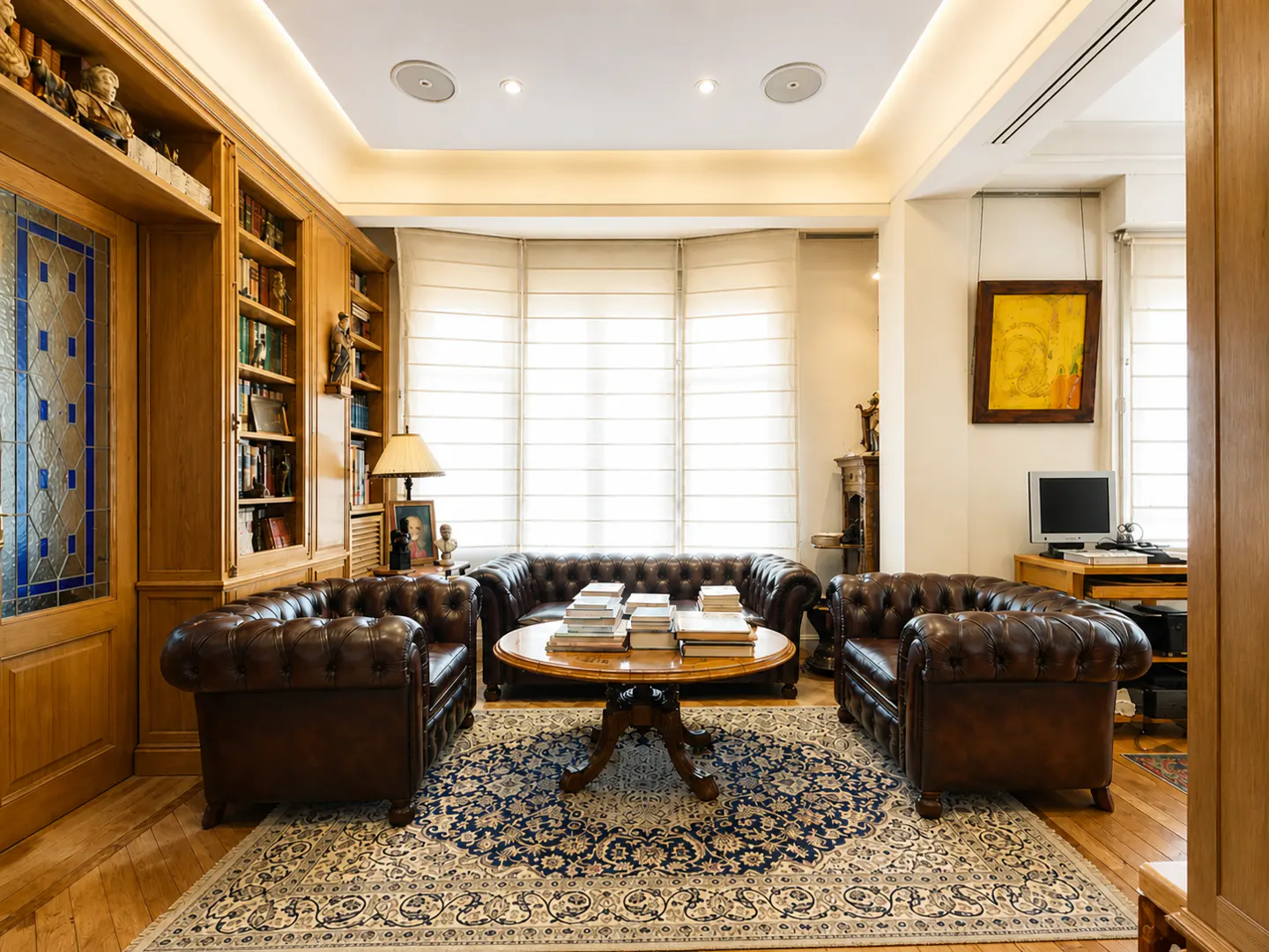 Living room with brown leather sofas, a round wooden table with books, and a blue patterned rug. Built-in wooden bookshelves line the wall.