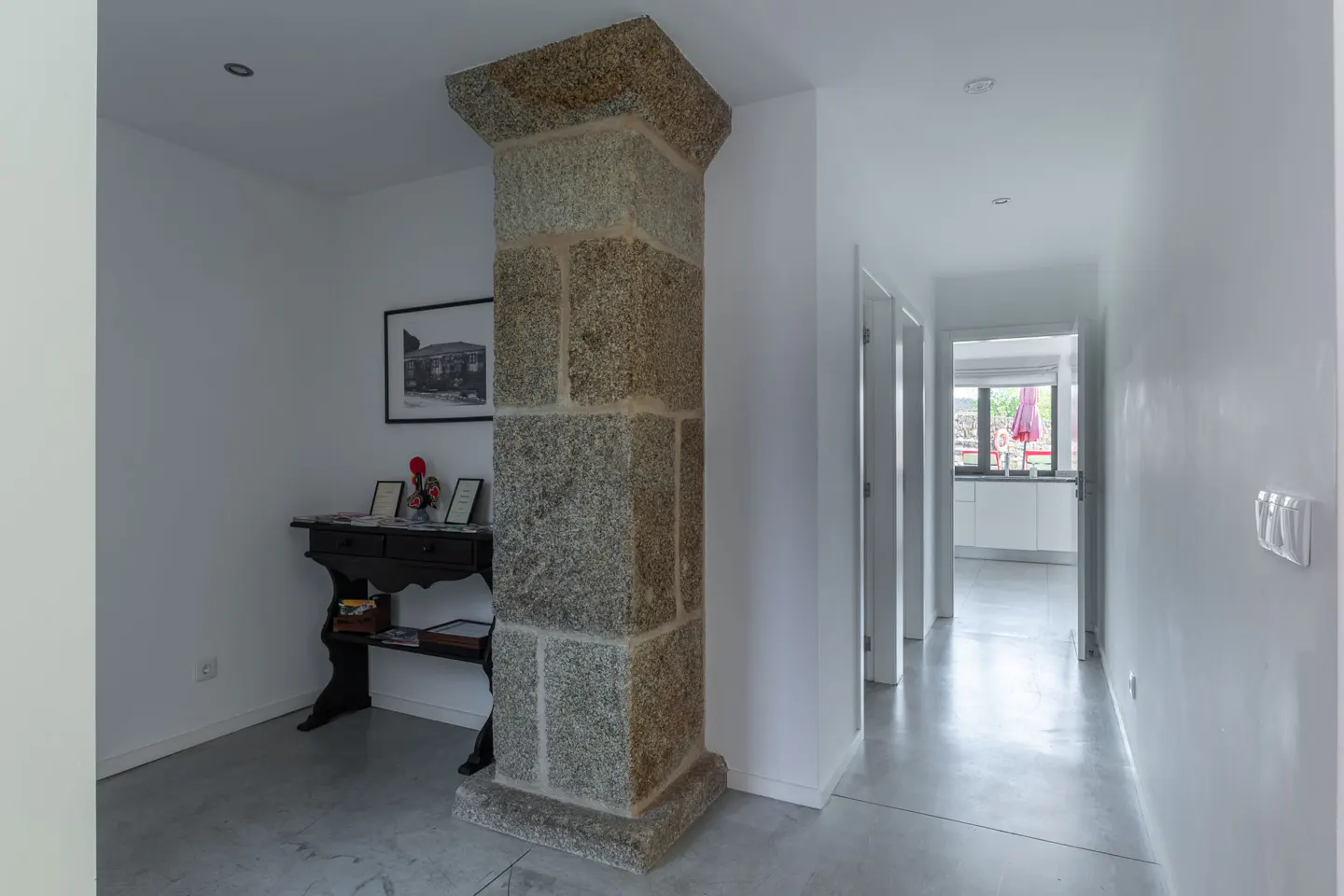 Interior view of a white hallway with a stone pillar, a black table with decorations, and a glimpse of a kitchen.