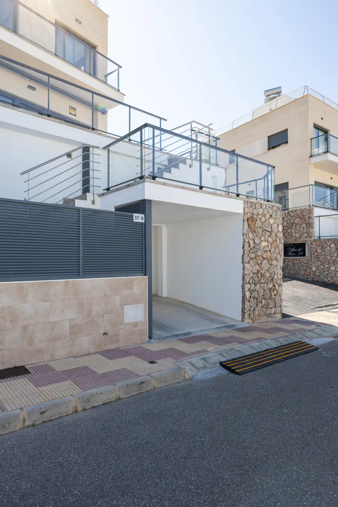 Exterior view of a modern, multi-story white building with a gray fence and stone accents. A covered parking area is visible.