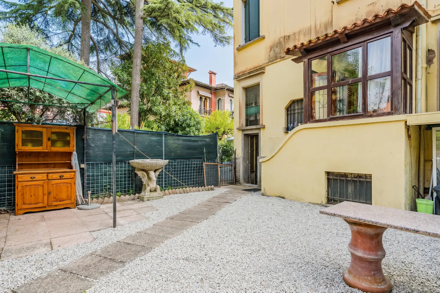 Outdoor patio with white gravel, stone path, and yellow building. A wooden hutch sits under a green awning, with a stone birdbath nearby.