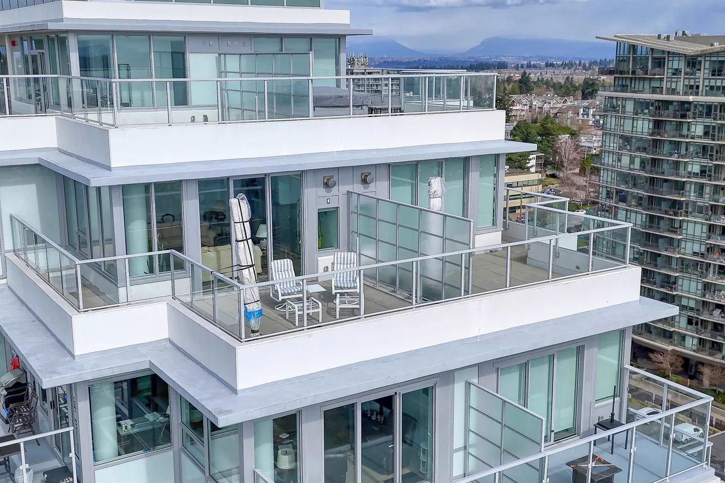 Condo building exterior with glass balconies. Two chairs and a table are on one balcony. Mountains are in the background.