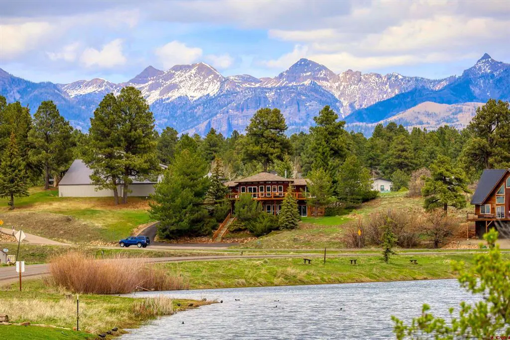 Scenic view of a lake with houses, trees, and snow-capped mountains in the background. A blue car is on the road.