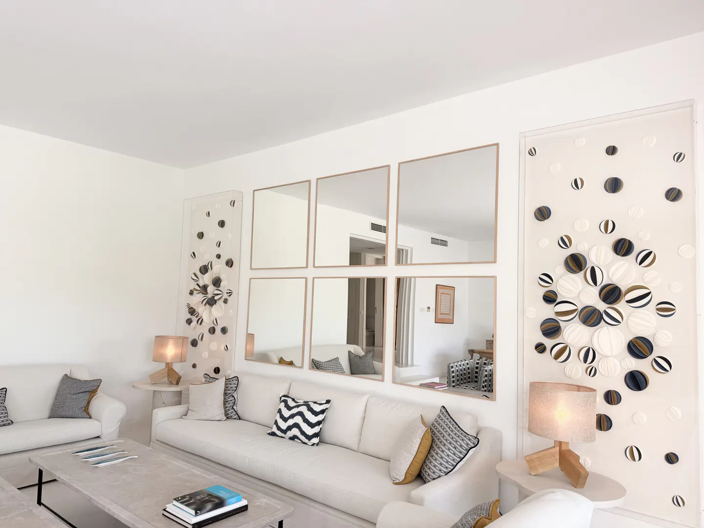 Bright living room with white sofas, patterned pillows, and a light gray coffee table. Six square mirrors and art panels decorate the white walls.