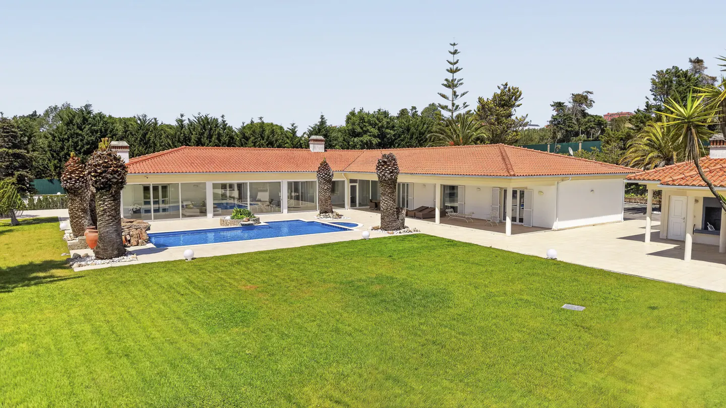 Single-story white villa with a red tile roof, large lawn, and a blue swimming pool surrounded by palm trees.