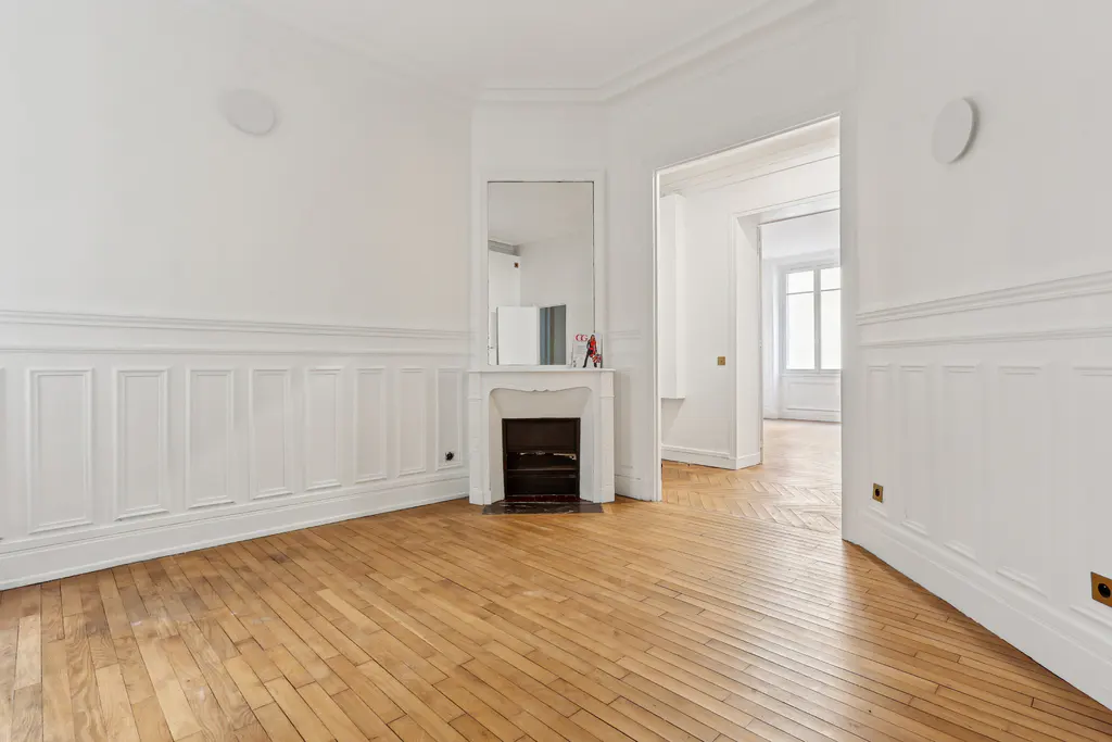 Empty room with herringbone wood floors, white walls with wainscoting, and a white fireplace with a mirror above it.