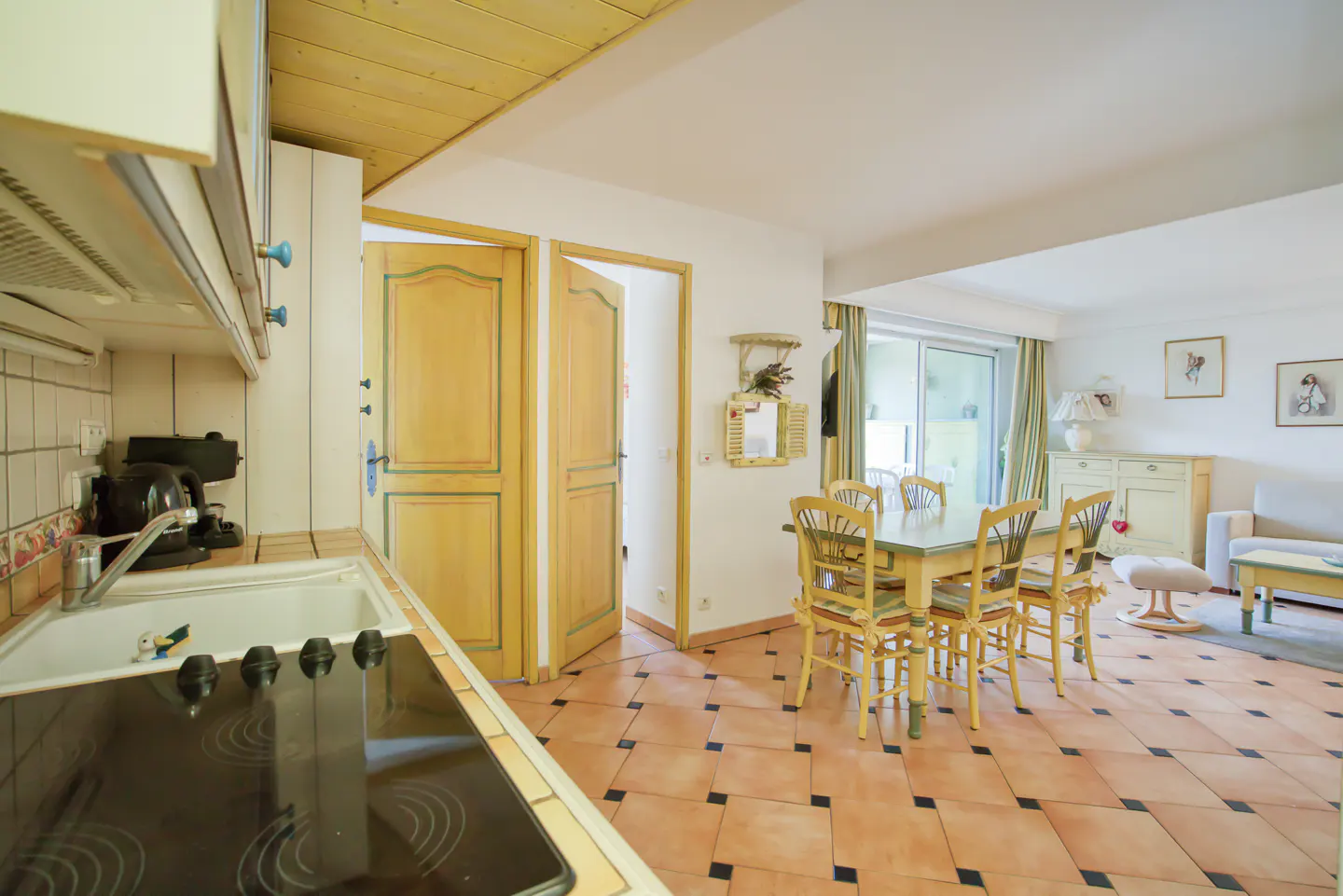 A bright kitchen and dining area with tile floors, wooden table and chairs, and a view into the living room.