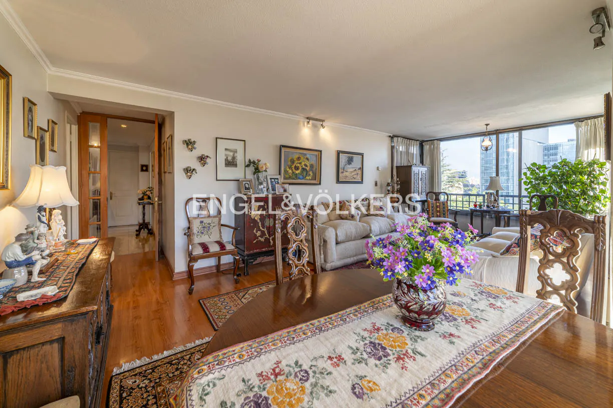 A bright living room with a wood table, floral centerpiece, and a view of the city through large windows. Engel & Völkers logo visible.