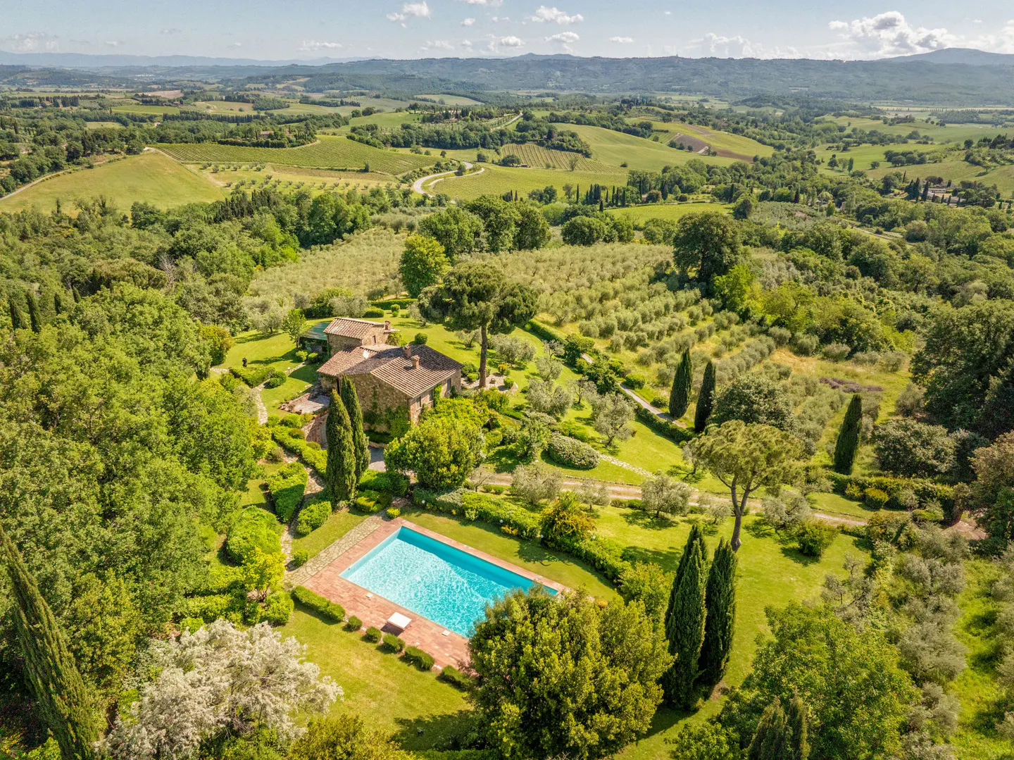 Aerial view of a Tuscan villa with a blue pool, surrounded by green trees and rolling hills.
