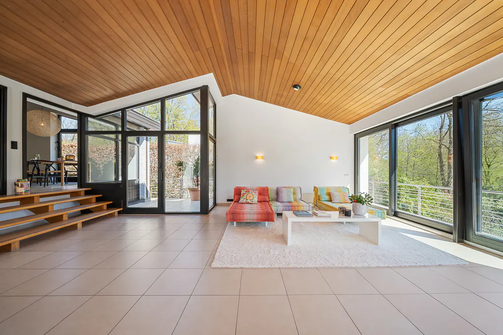 Bright living room with wood ceiling, tile floor, and large windows. Colorful couches sit on a white rug near a white coffee table.