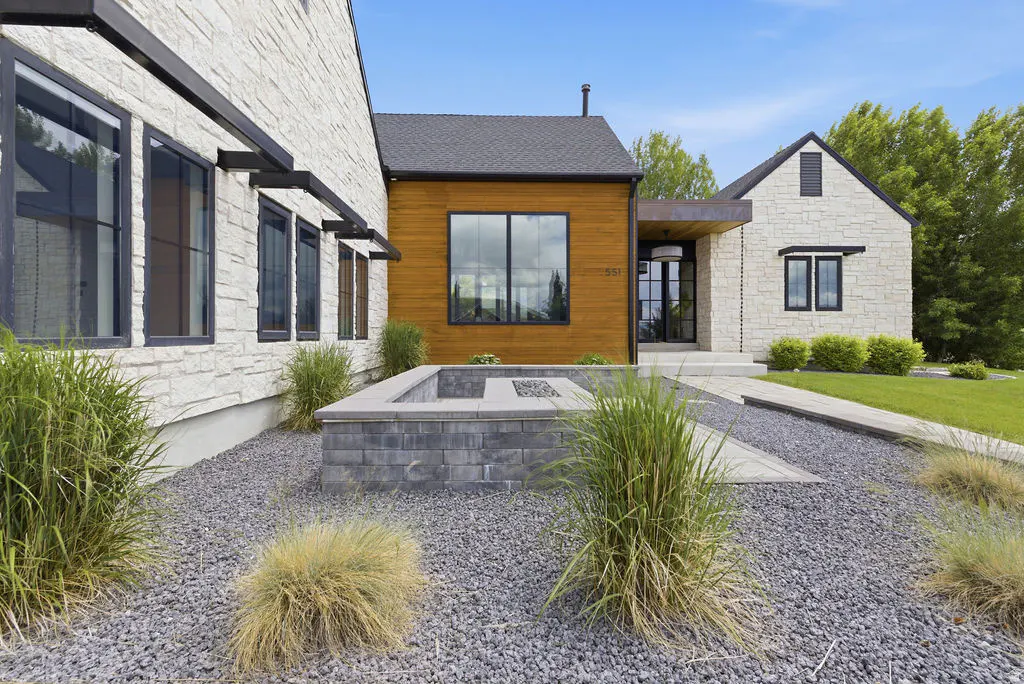 Exterior of a modern home with stone and wood siding, black framed windows, and a stone fire pit in the front yard.