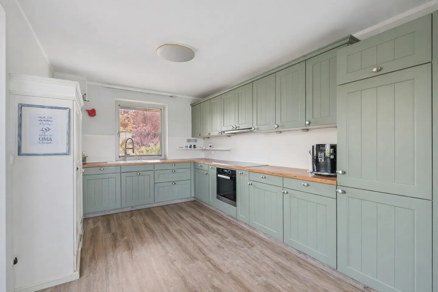 A kitchen with light green cabinets, wood countertops, and wood-look flooring. A window is above the sink.