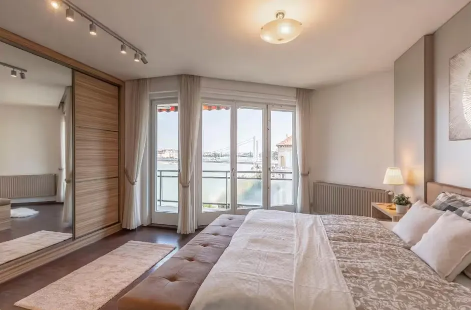 Modern bedroom with a large bed, mirrored wardrobe, and glass doors opening to a balcony with a river view.