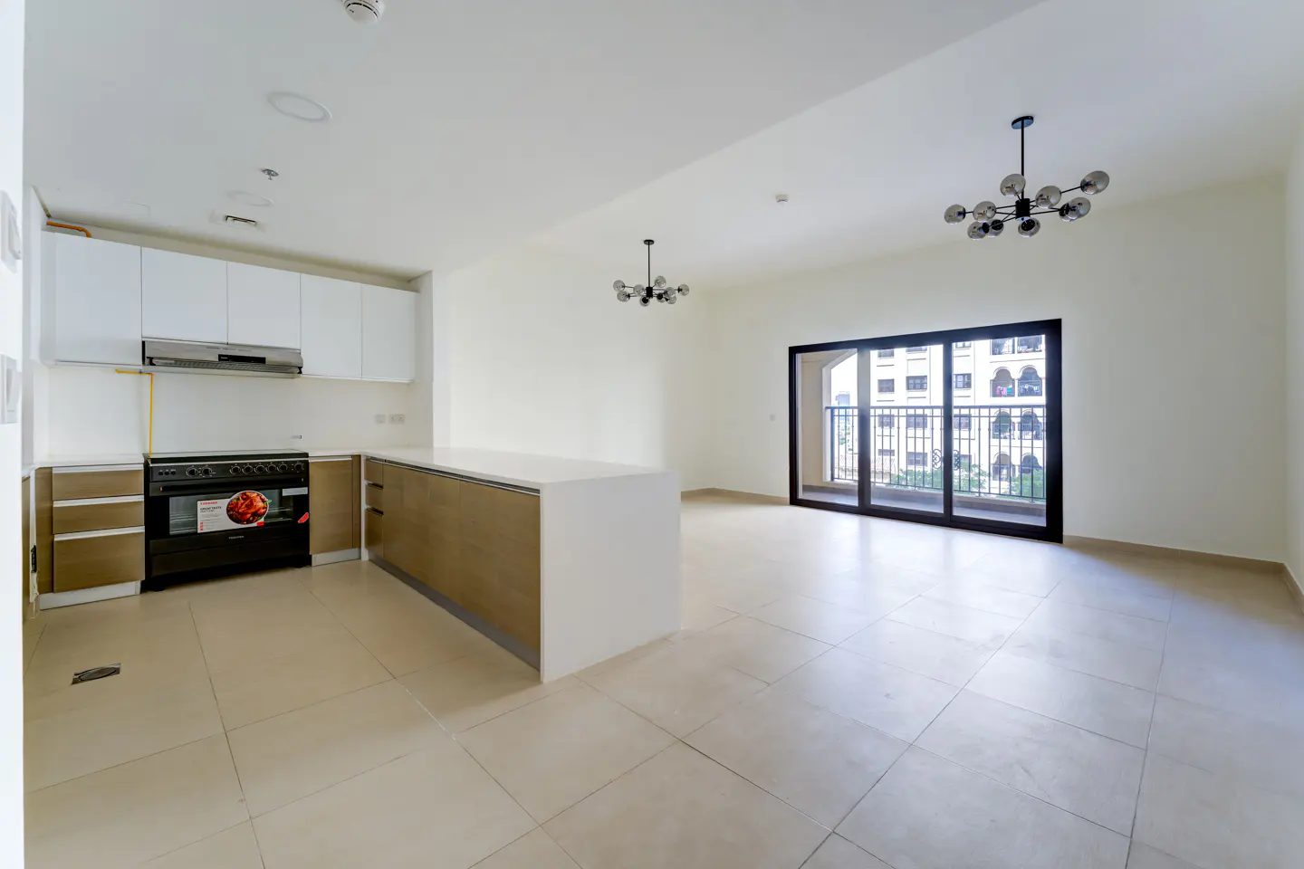 Bright, empty apartment with white walls, tile floors, and a kitchen with white cabinets and a black oven. A sliding glass door leads to a balcony.