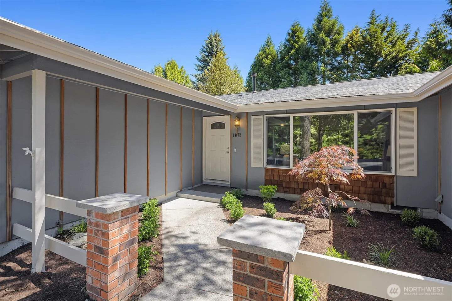 Exterior of a gray house with a white door, large window, and brick accents. A concrete walkway leads to the entrance.