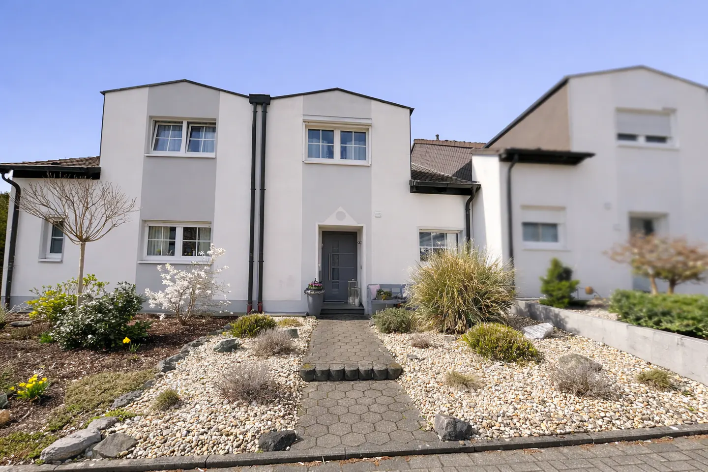 Exterior view of a two-story white and gray house with a stone walkway leading to a gray front door. The yard is landscaped with rocks and greenery.