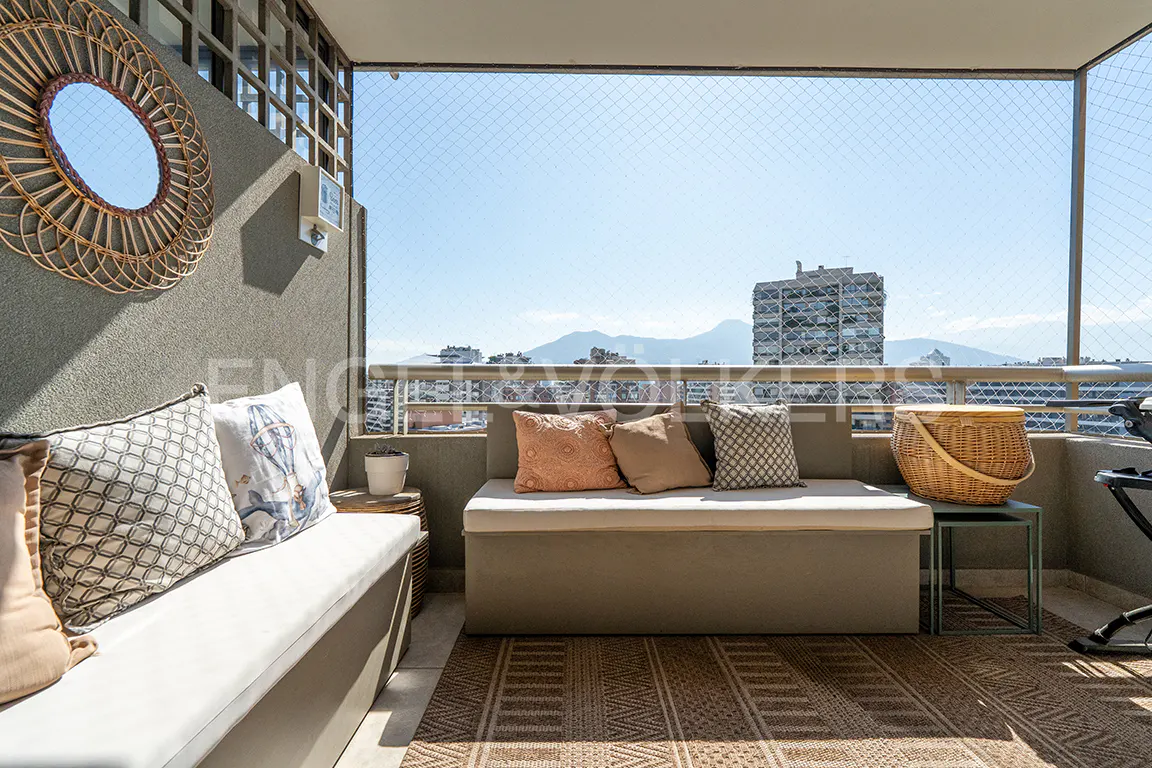 Balcony view with seating, pillows, and a wicker mirror on a gray wall. Cityscape and mountains visible through a safety net.
