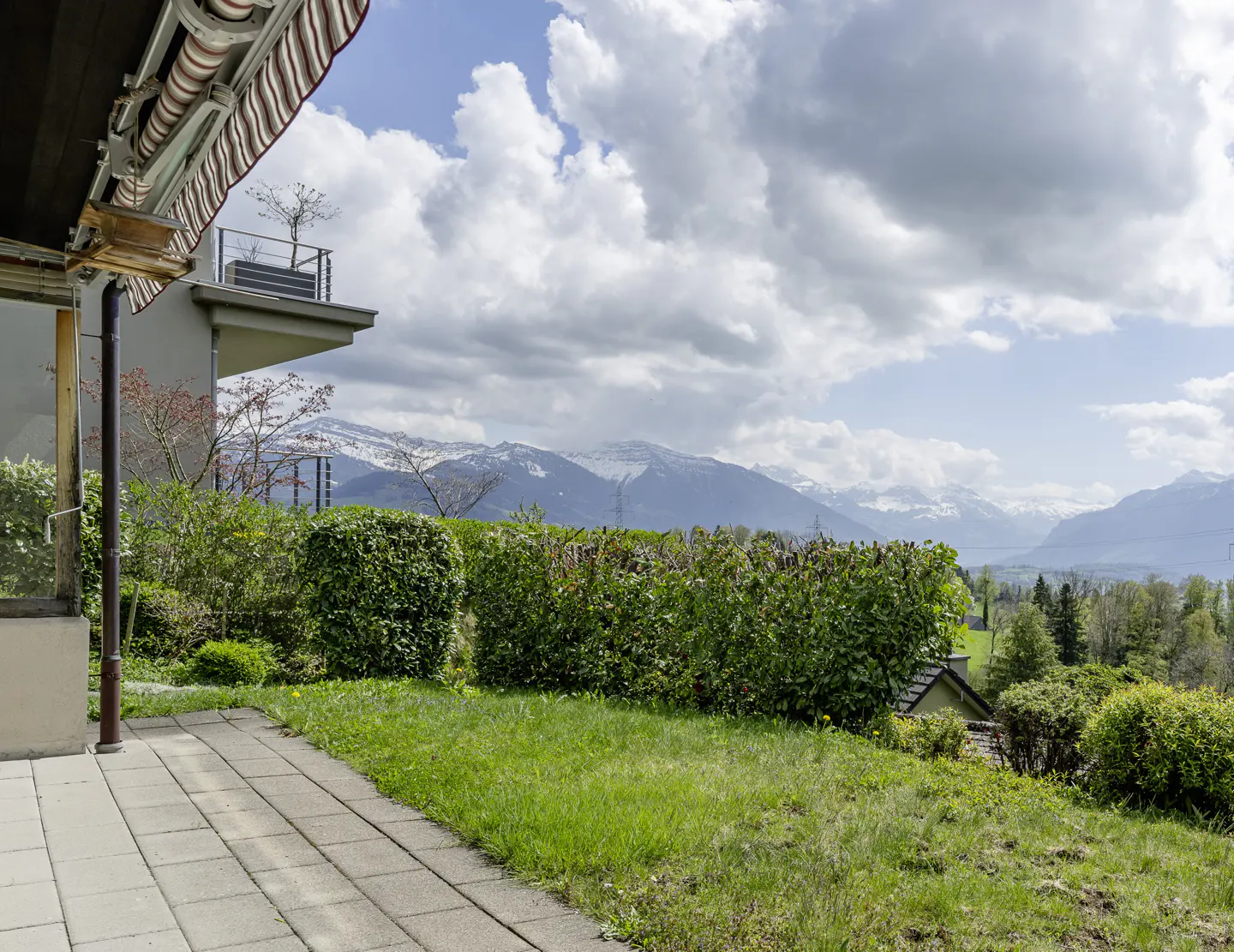 View from a patio with green grass and bushes, overlooking snow-capped mountains under a cloudy sky.