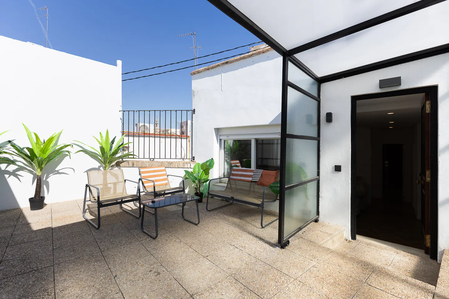 Outdoor patio with seating, plants, and a black-framed glass enclosure leading to an open doorway. White walls and blue sky in the background.