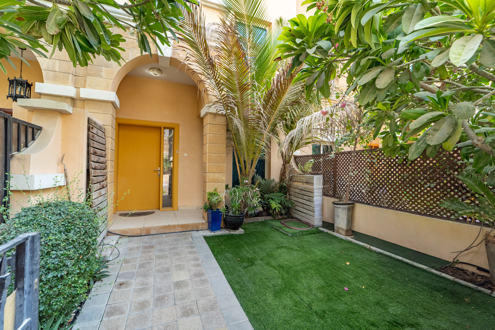 A bright yellow front door set in a tan arched entryway, with a stone path leading to a small green artificial lawn.