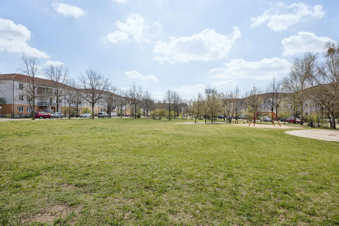 A wide green lawn with trees and buildings under a blue sky with white clouds. A playground is visible in the background.